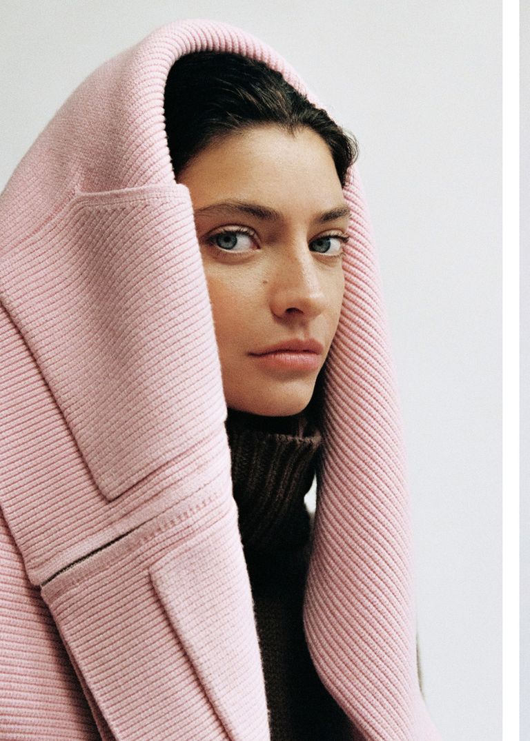 Woman wearing a pink ribbed coat with the hood up, over a brown sweater, gazing to the side against a neutral background.