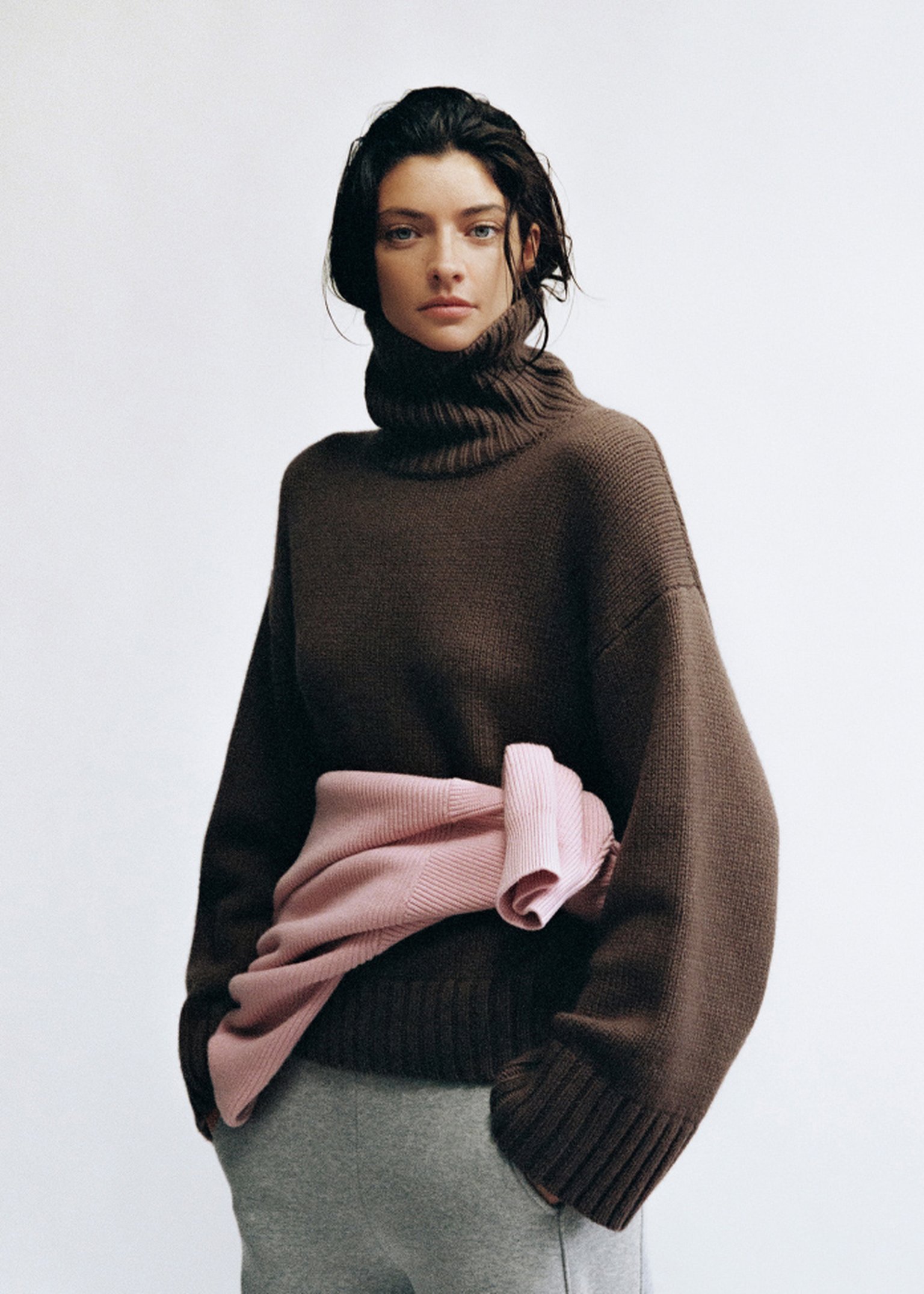 Woman in a brown turtleneck sweater, pink sweater tied around her waist, and gray pants stands against a plain background.