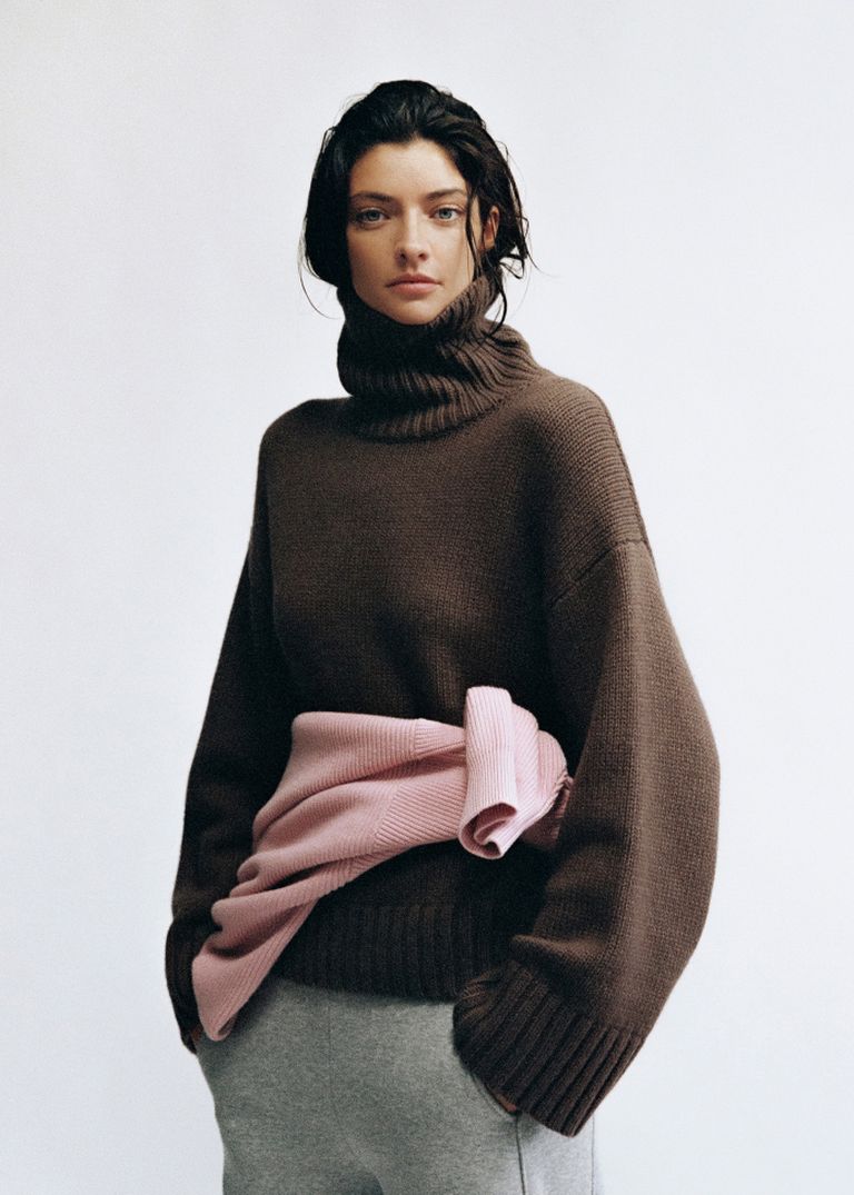 Woman in a brown turtleneck sweater, pink sweater tied around her waist, and gray pants stands against a plain background.