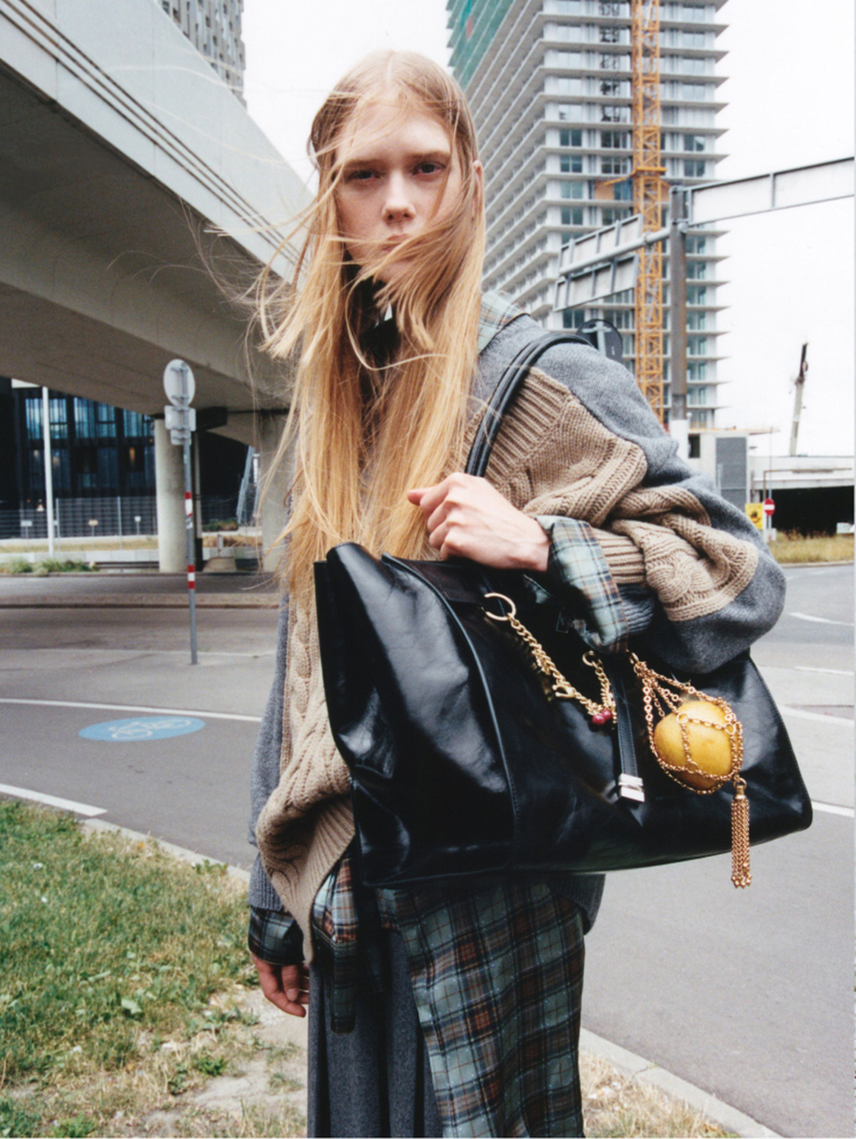 Young woman with long hair stands outdoors, wearing layered clothing and holding a large black bag, with urban buildings in the background.