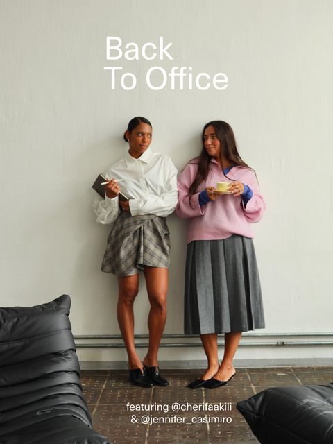 Two people stand against a wall in office attire, one in a white shirt and plaid skirt, the other in a pink sweater and grey skirt, holding cups.