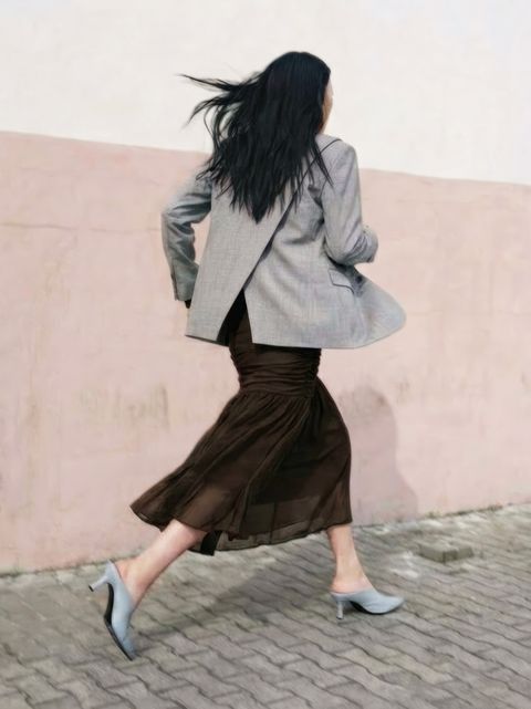 A woman in a grey blazer and brown skirt walks briskly in high heels on a cobblestone path against a pale pink wall.