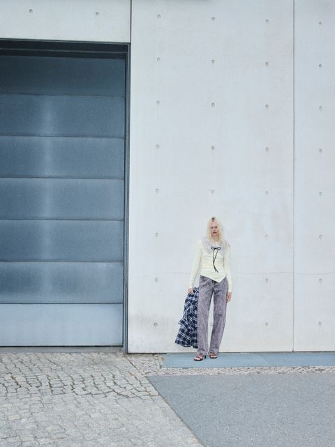 A person stands against a large concrete wall, holding a plaid jacket, wearing a light top and pants. The setting appears urban and minimalist.