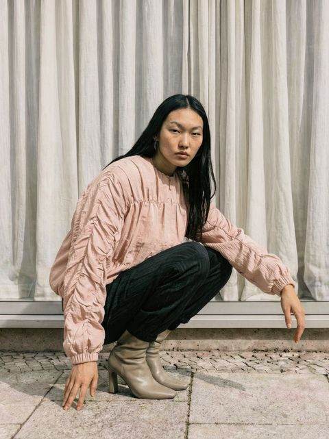 A person squatting outdoors against a curtain backdrop, wearing a pink blouse, black pants, and beige ankle boots, with long dark hair and hoop earrings.