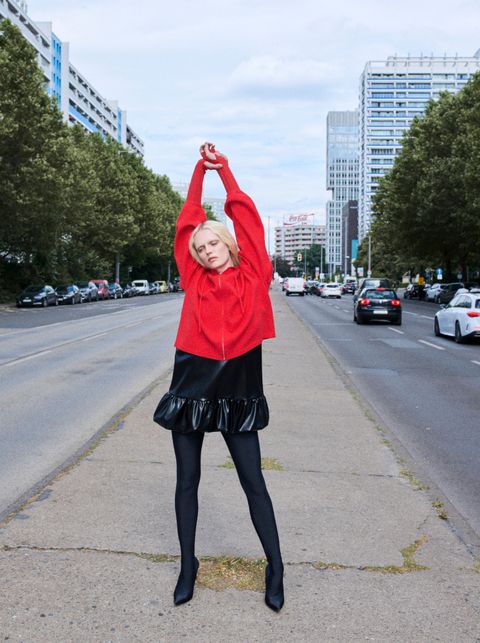 Person in a red top and black skirt stands with arms raised on an urban street, with buildings and trees in the background.