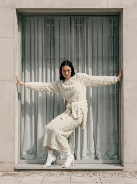 Person in a textured white outfit poses confidently in a large window frame, with arms outstretched and wearing white high-heeled sandals.