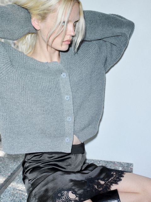 A blonde woman in a gray buttoned sweater and black lace-trimmed skirt sits casually with hands in her hair, looking to the side.