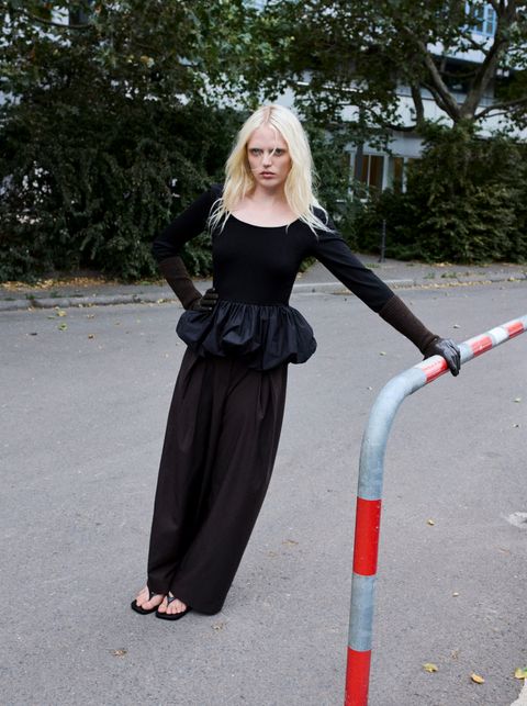 A person with long blonde hair leans against a red and white barrier. They wear a black outfit with a ruffled top and wide-leg pants. Trees in the background.