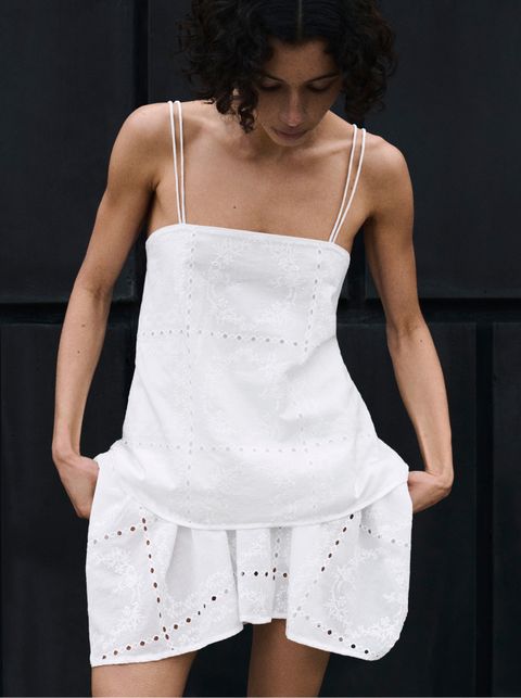 A woman in a white dress with thin straps and floral embroidery stands against a dark background, looking down with hands in pockets.