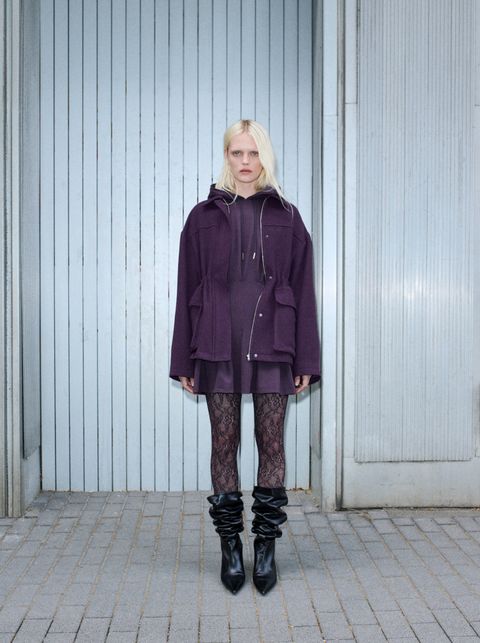 Person in a purple coat, lace leggings, and black boots, standing against a corrugated metal wall.