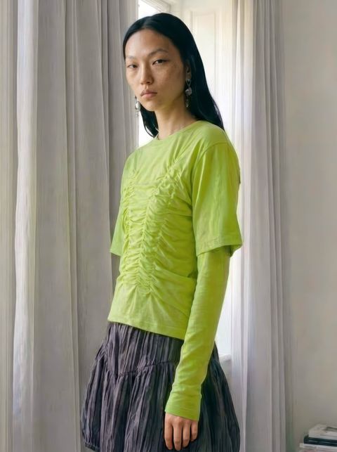 A person with long black hair wearing a lime green ruched top and gray skirt stands in front of white curtains.