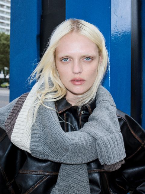 Person with long blonde hair, wearing a black leather jacket and a thick gray scarf, standing in front of a blue wall outdoors.