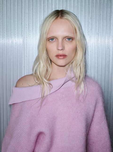 A person with long blonde hair wearing a pink off-the-shoulder sweater stands against a textured metallic background.