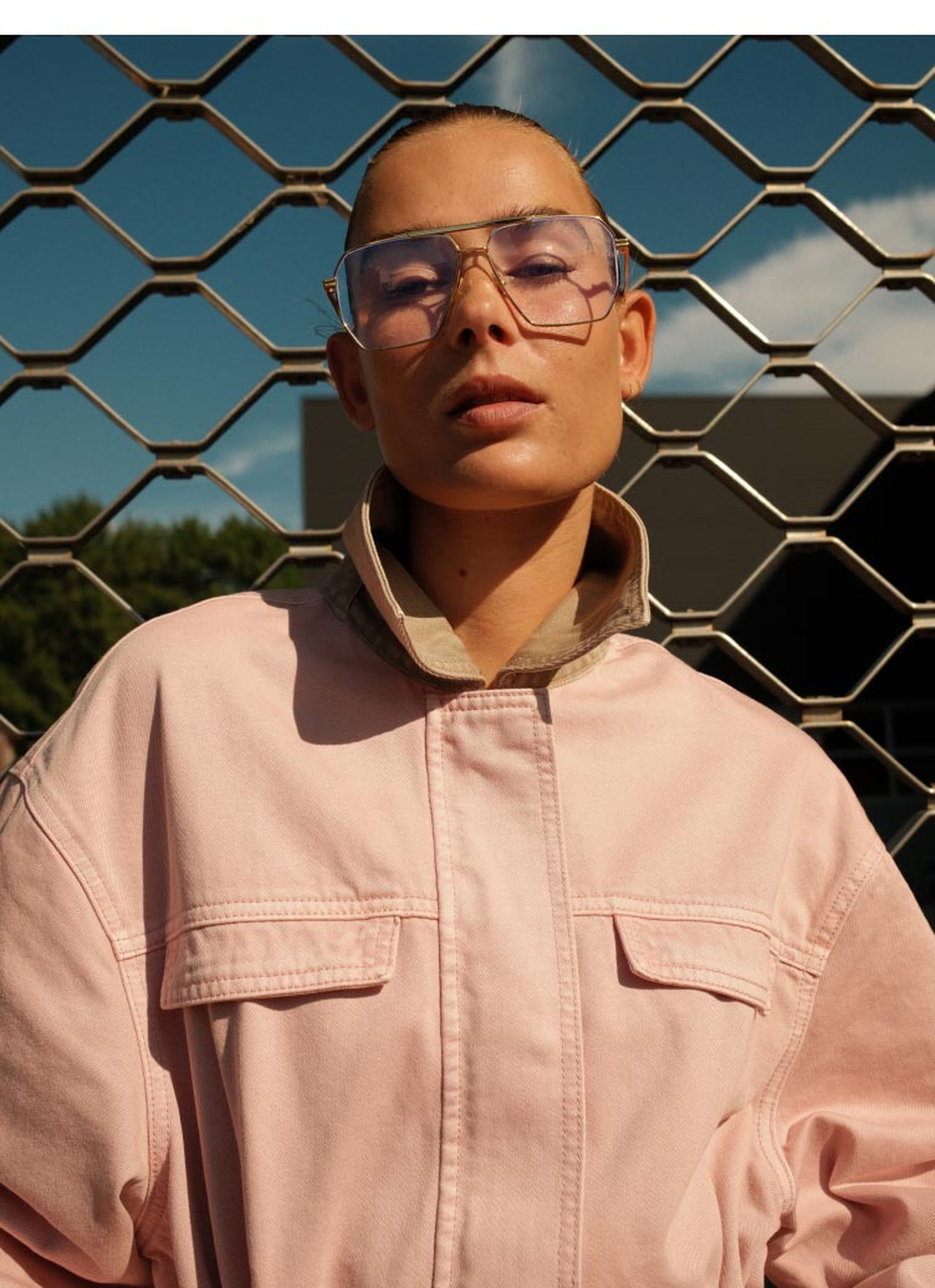 Person in a pink jacket and large clear-rimmed glasses poses confidently against a chain-link fence, with a blue sky in the background.