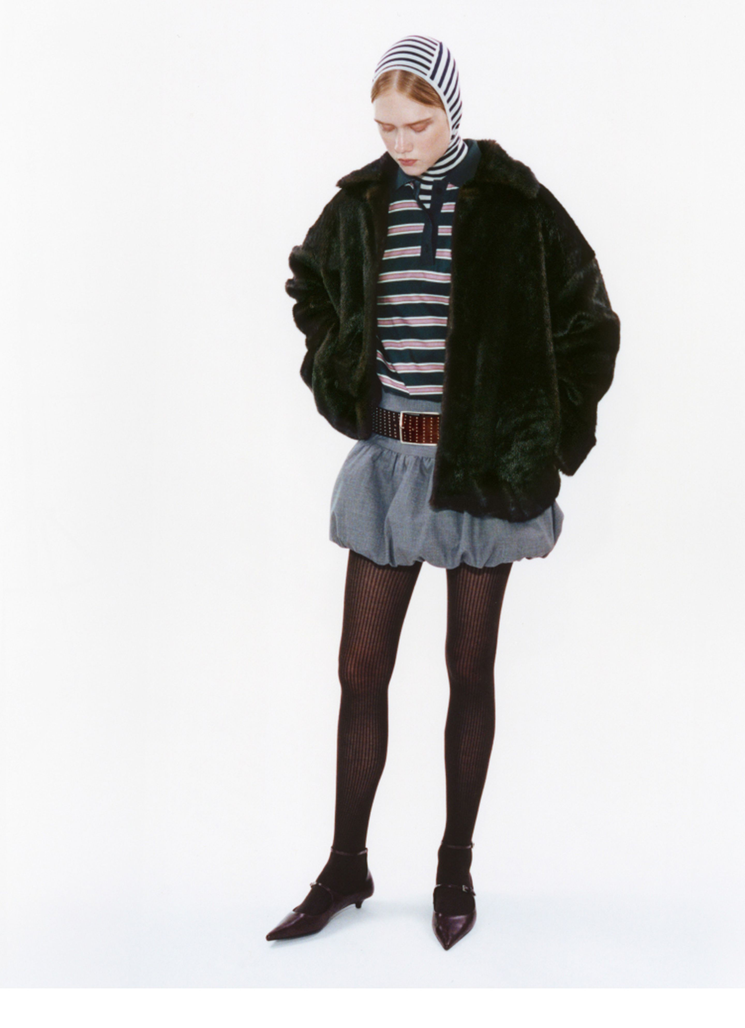 Person wearing a striped top, dark jacket, gray skirt, striped headscarf, and black tights with pointed shoes, standing against a white background.