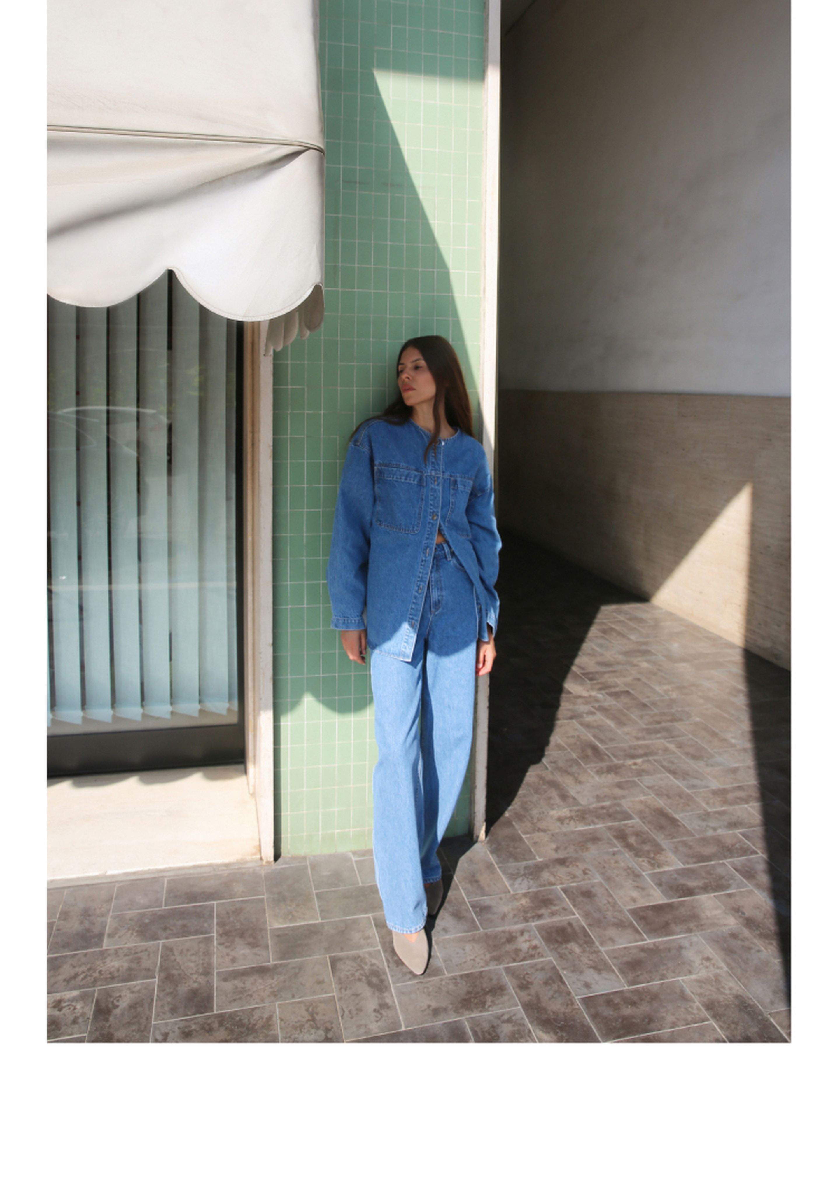 Woman in denim jacket and jeans stands against a green tiled wall, with sunlight casting shadows on the tiled floor.