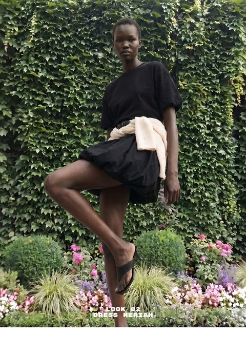 Person in a black dress with a beige sweater tied at the waist stands in front of a lush green vine wall and a colorful flower garden.