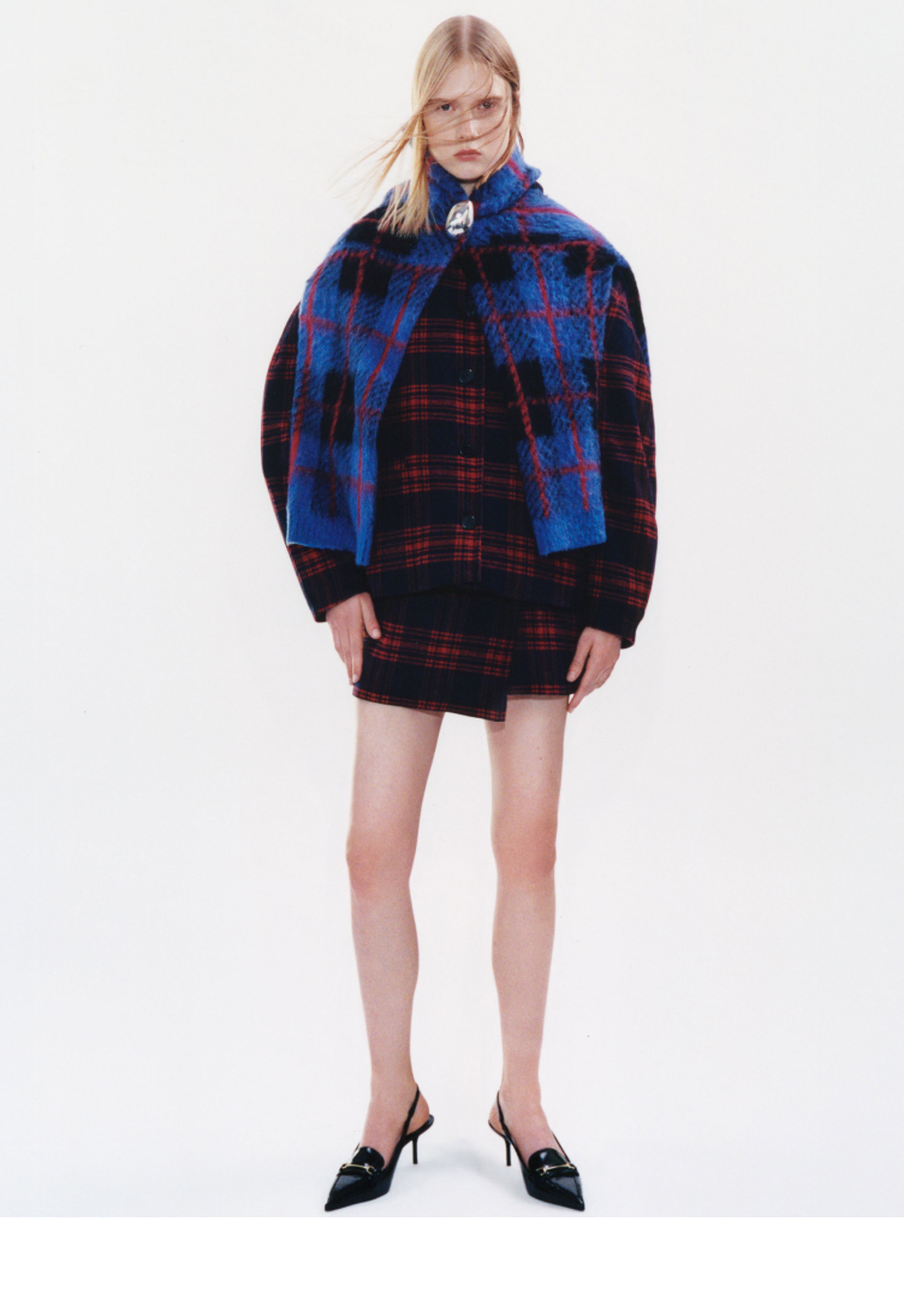 Person wearing a plaid outfit with a blue and red pattern, black heels, and glasses, standing against a plain background.