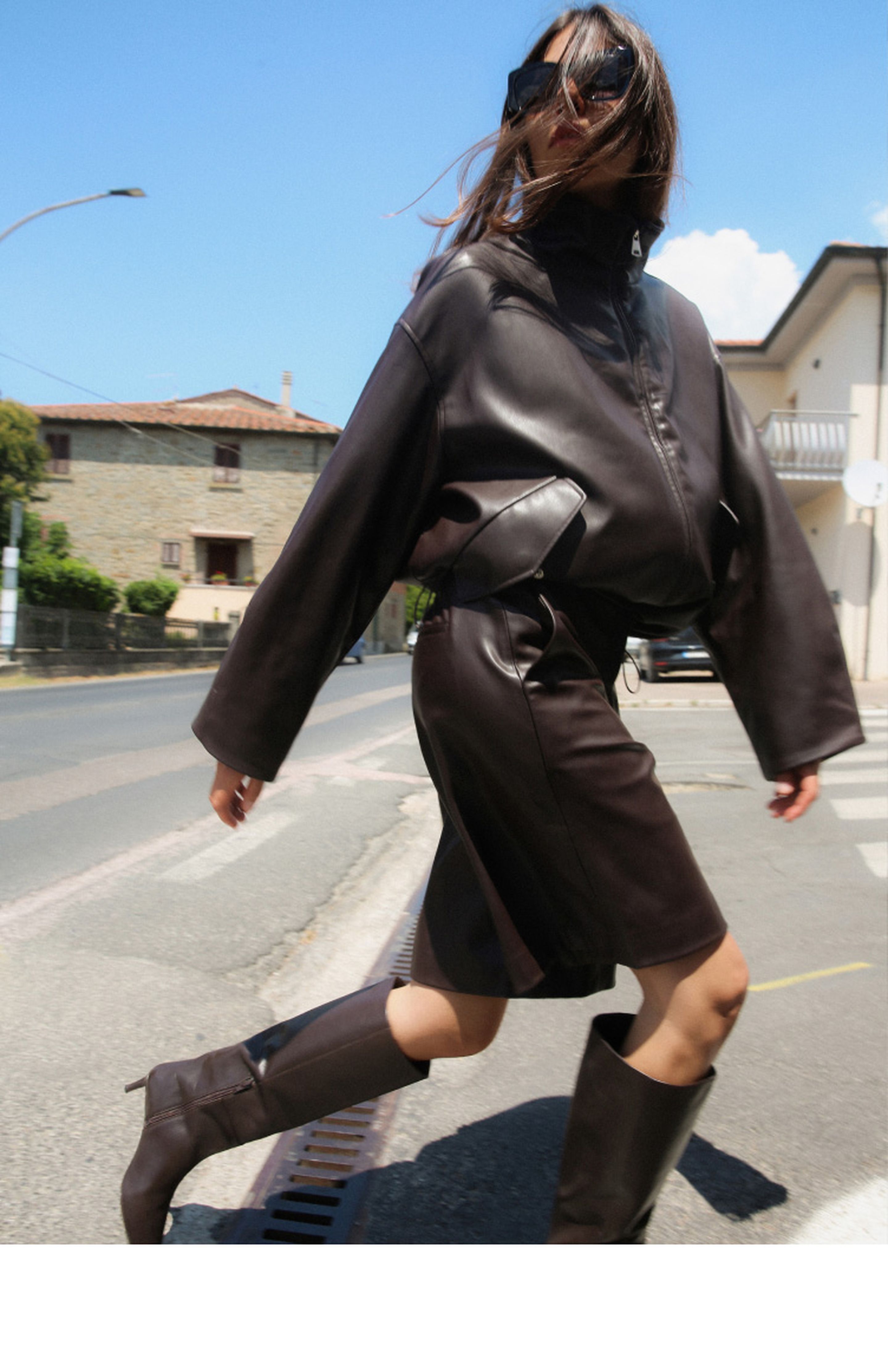 Person in dark leather outfit and boots walks energetically on a sunny street, with buildings and a clear blue sky in the background.