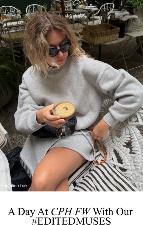 Person in a gray outfit and sunglasses holding a coffee cocktail, sitting at an outdoor cafe with wicker chairs and a table setting.