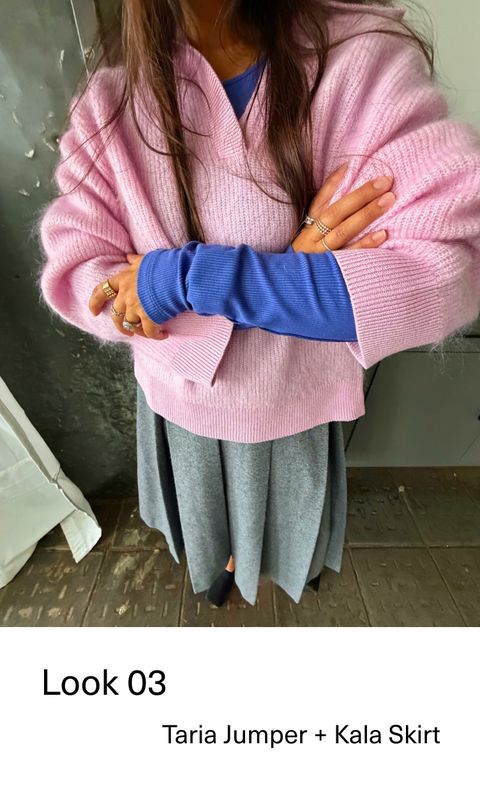 Person wearing a pink jumper over a blue top, paired with a gray skirt, standing with arms crossed.