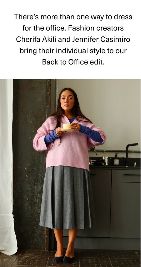 A woman stands in an office kitchen wearing a pink sweater over a blue shirt with a gray skirt, holding a cup and saucer, styled for work.
