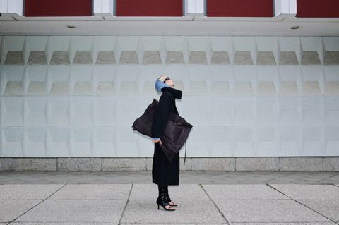 A person in a black coat and boots stands on a patterned sidewalk, looking up with a large tote bag against a textured wall.