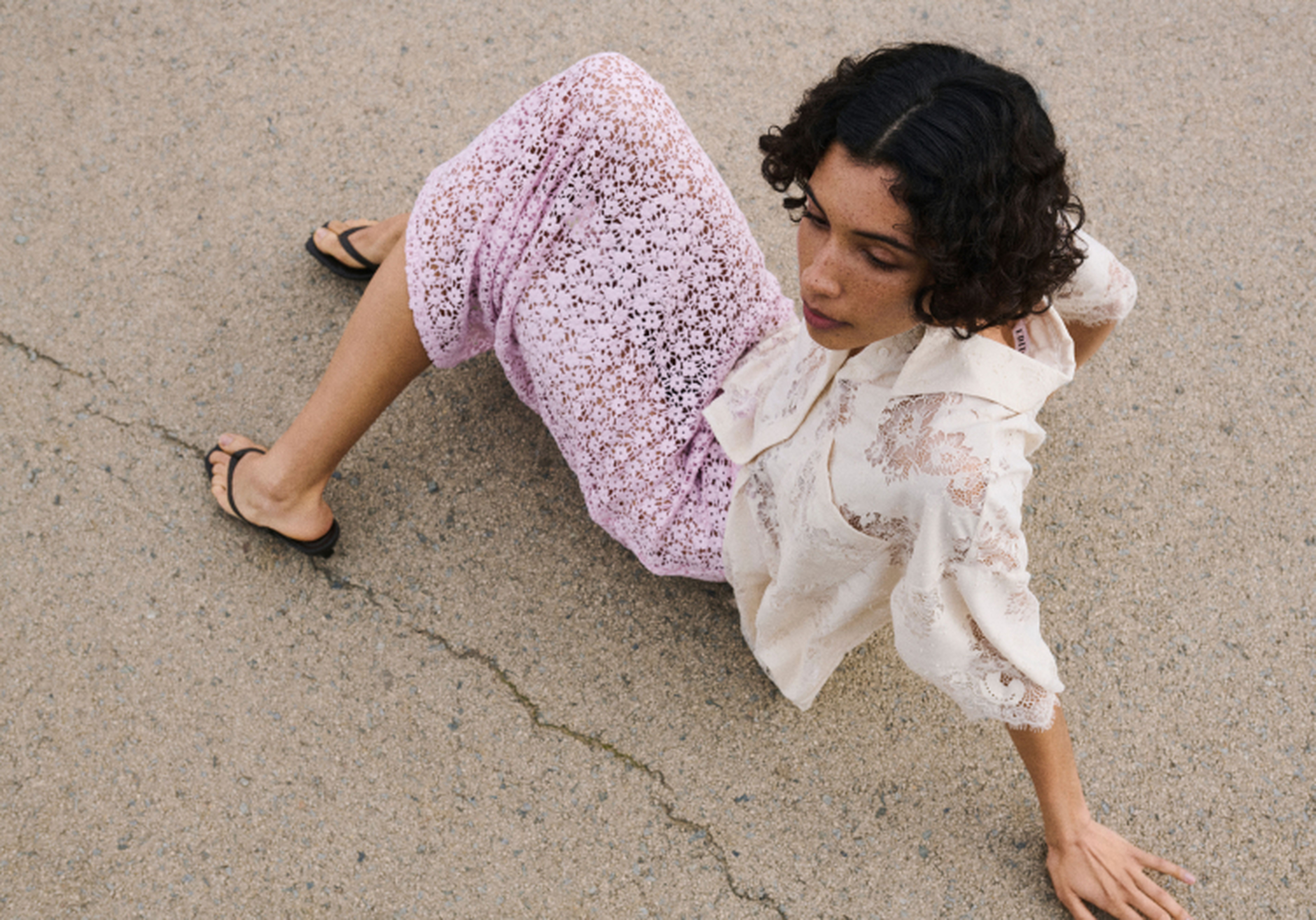 Person in a lace outfit sitting on the ground, looking to the side. The background is a concrete surface with a visible crack.