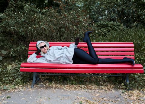 A person in a gray hoodie and black pants lounges on a red bench in a park, surrounded by green foliage and fallen leaves.