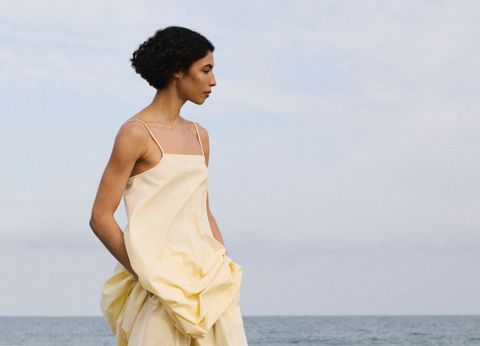 Woman in a flowing yellow dress stands by the ocean, gazing into the distance under a cloudy sky.