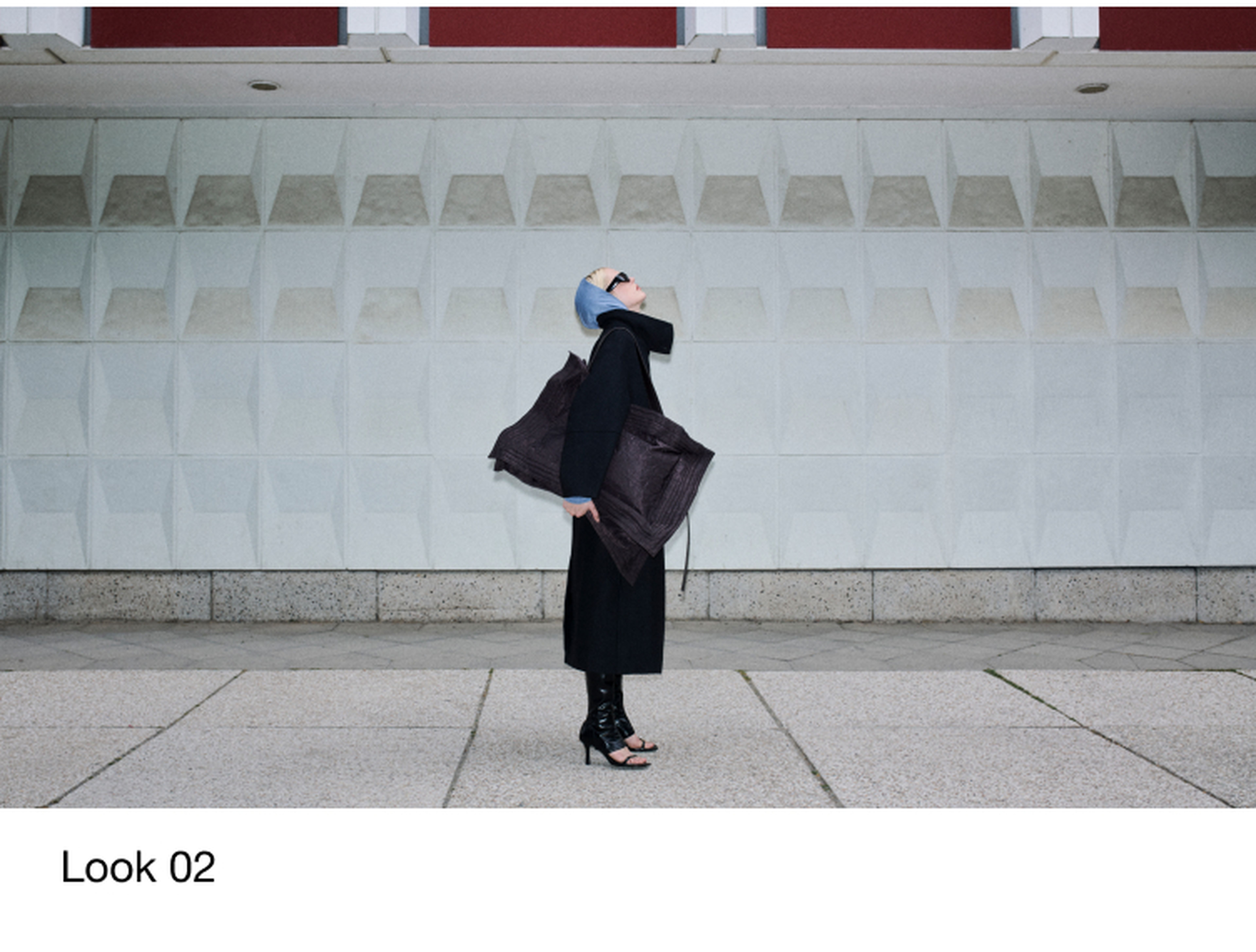A person in a black coat, oversized bag, and heeled sandals stands against a textured wall, looking upward, wearing a blue headscarf and sunglasses.