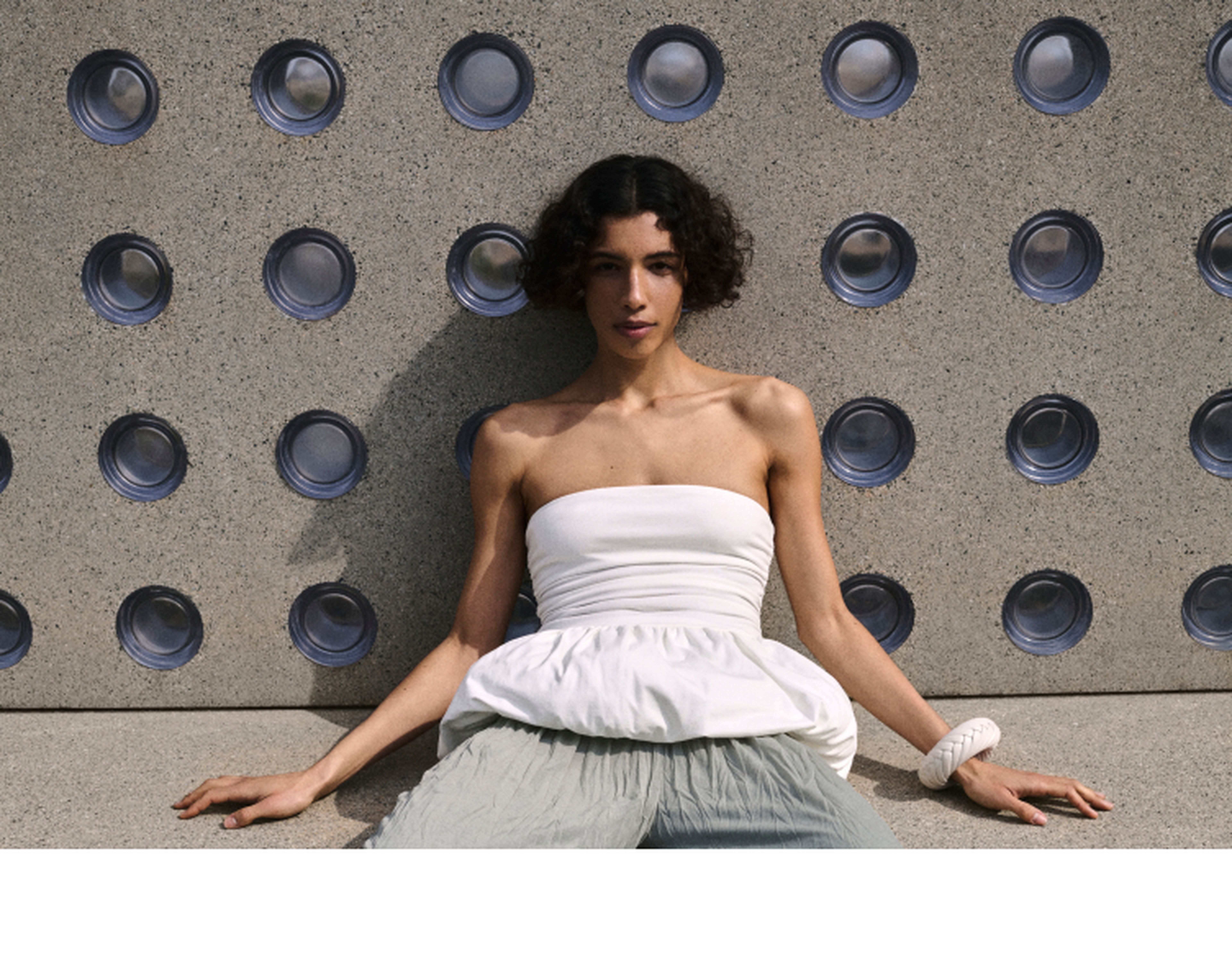 Person in a white strapless top and light pants sitting against a textured wall with circular patterns.