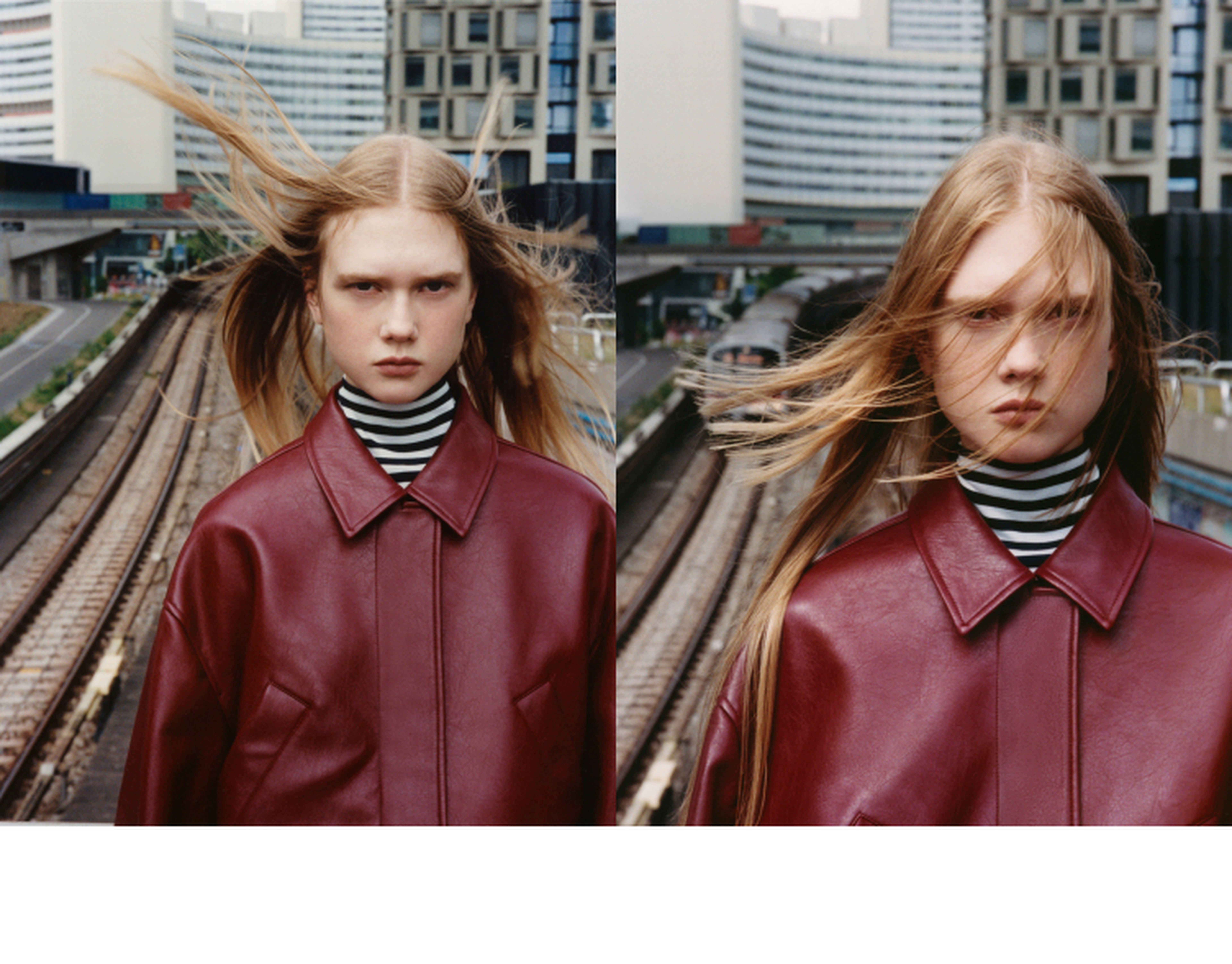 Person with long hair and a red leather jacket stands on a railway platform, with wind blowing their hair, against an urban backdrop.