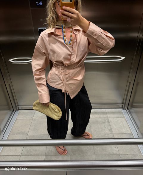 Woman in pink wrap blouse and black pants taking a mirror selfie in an elevator, holding a yellow clutch, with beaded necklace.