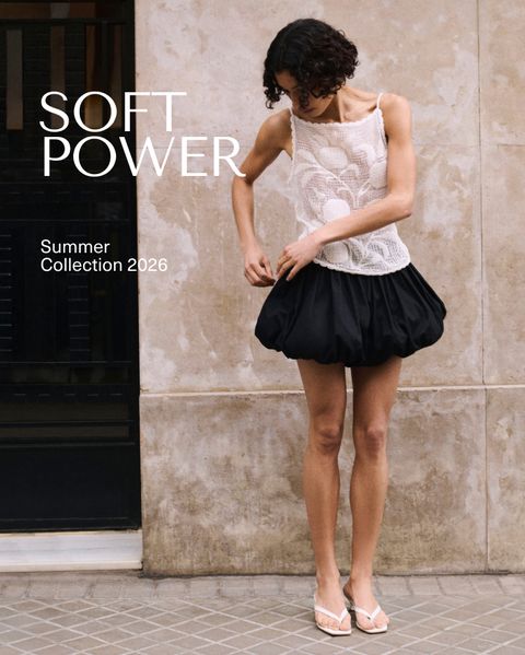 Model in a white crochet top and black skirt stands against a stone wall, showcasing "Soft Power" Summer Collection 2026.
