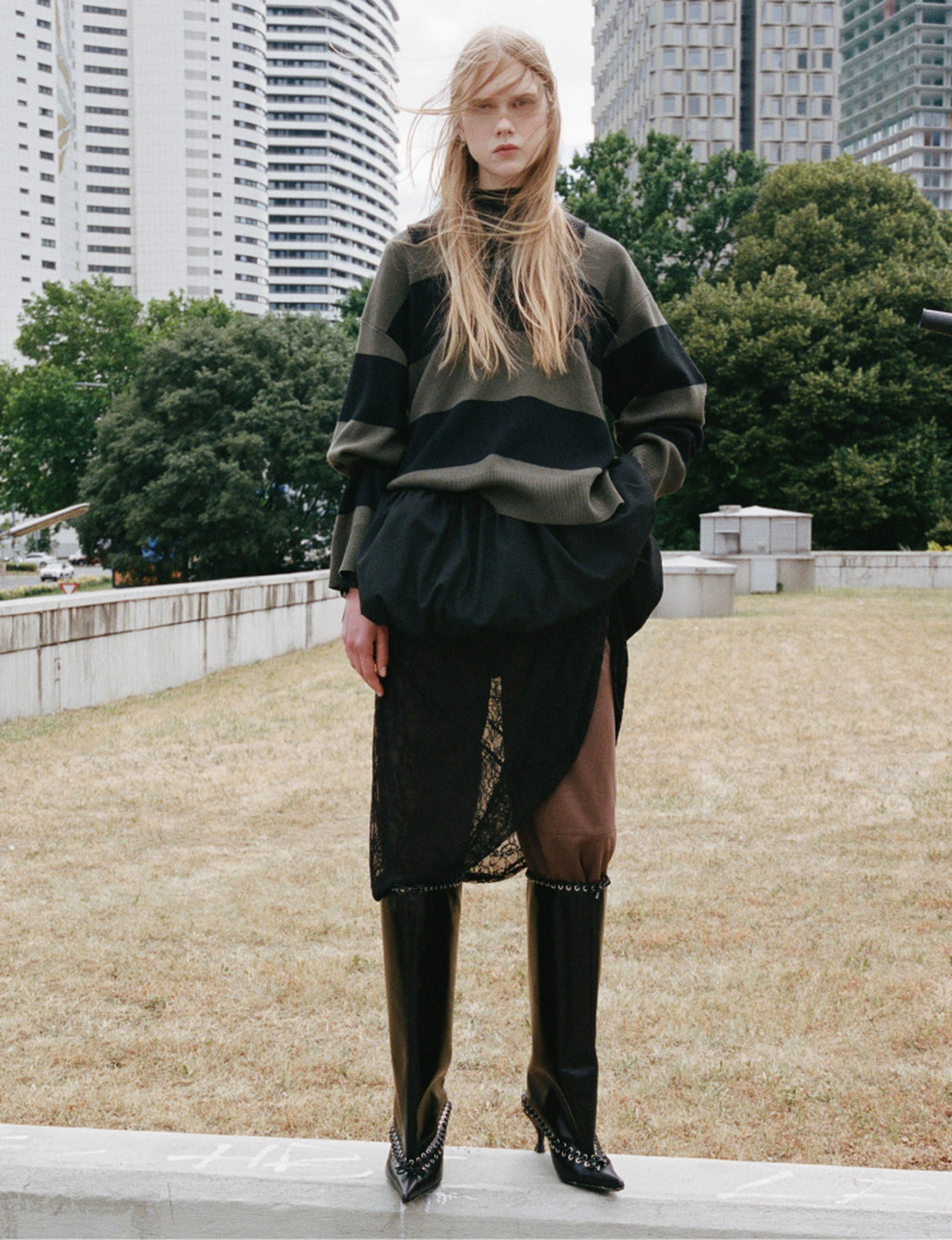 A person stands outdoors in a layered outfit with a striped sweater and tall boots. Buildings and trees are visible in the background.