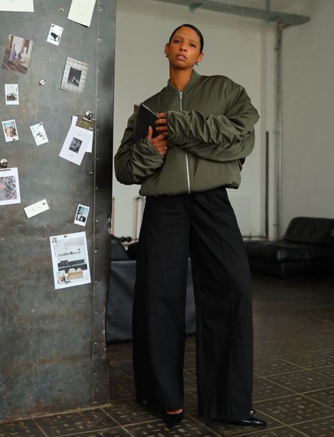 A person in a green bomber jacket and black wide-leg pants holds a book, stands by a wall with various photos and notes in a modern interior room.