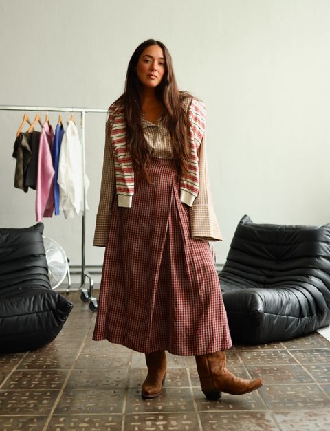A woman with long hair wearing a checked ensemble and boots stands in a room with clothing on a rack and black cushioned chairs.