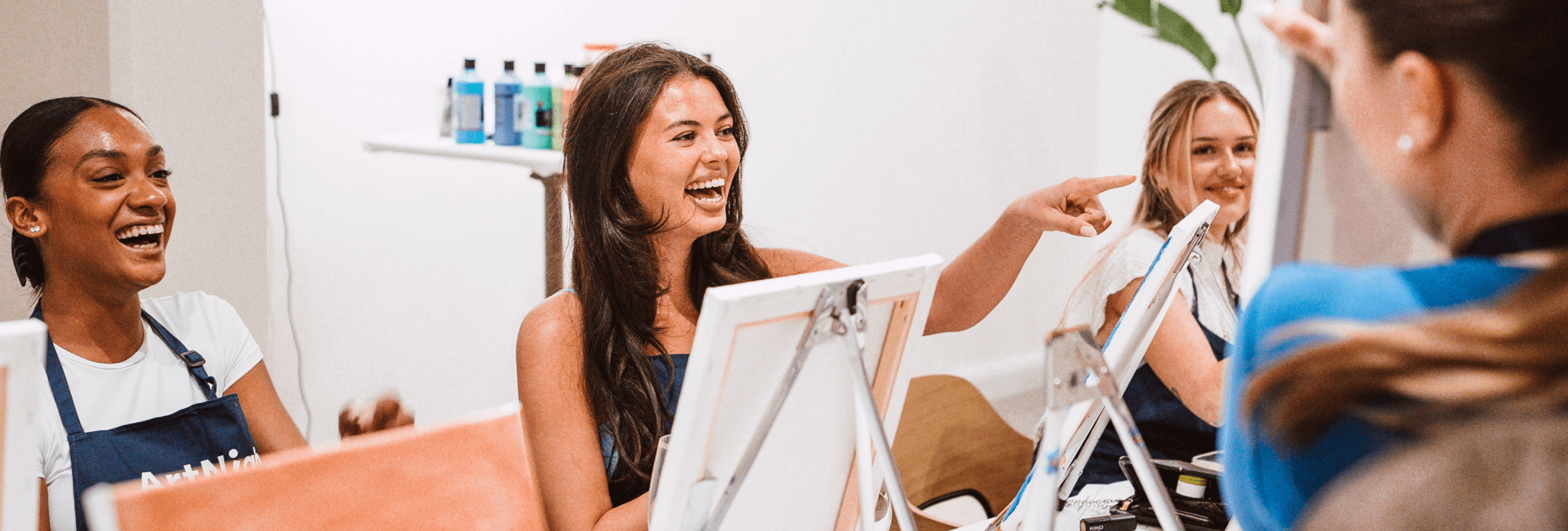 Menschen, die an einem Malkurs teilnehmen, an einem Tisch mit Staffeleien, Leinwänden und Getränken sitzen, lächeln und sich miteinander unterhalten.