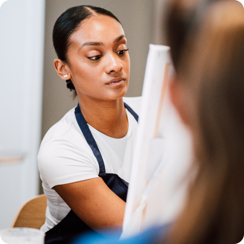 Frau in weißem Hemd und blauer Schürze konzentriert sich auf das Malen an einer Staffelei in einem gut beleuchteten Raum.