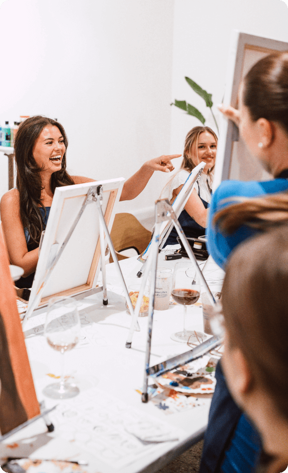 Menschen, die an einem Malkurs teilnehmen, an einem Tisch mit Staffeleien, Leinwänden und Getränken sitzen, lächeln und sich miteinander unterhalten.