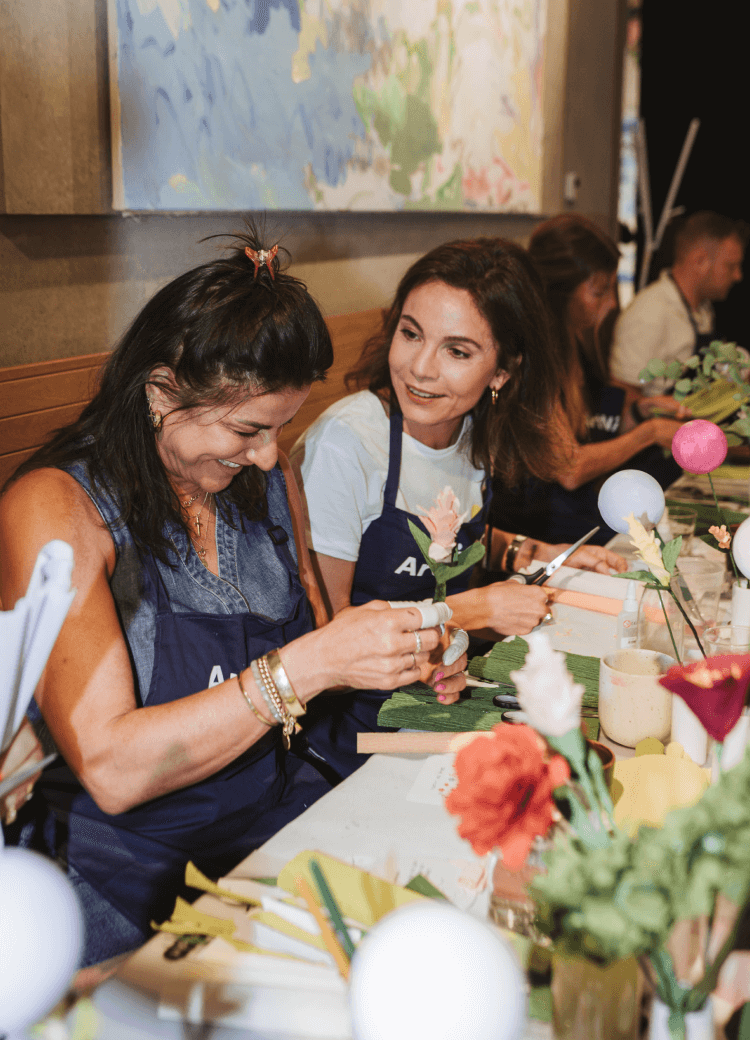 Zwei Frauen mit Schürzen lächeln, während sie an einem Tisch mit Blumen basteln. Andere sind im Hintergrund mit ähnlichen Tätigkeiten beschäftigt.