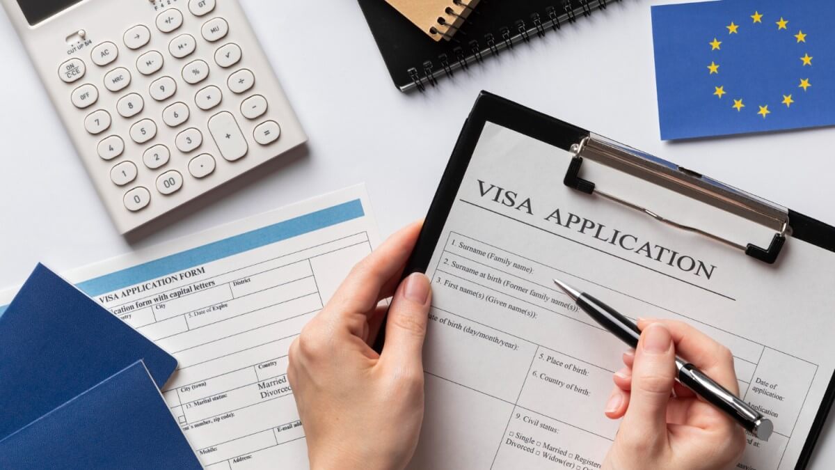 Các loại Visa phổ biến bạn cần biết Hands filling out a visa application form on a clipboard, with a calculator, notepads, and an EU flag in the background.