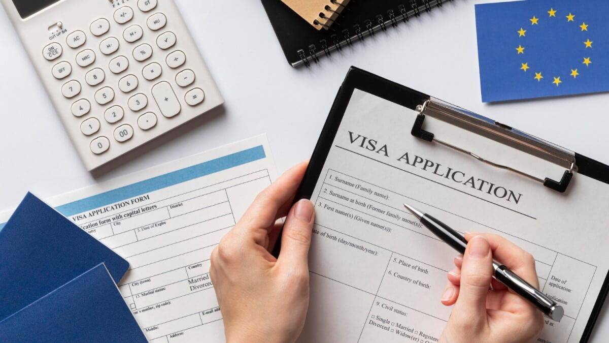 Hands filling out a visa application form on a clipboard, with a calculator, notepads, and an EU flag in the background.