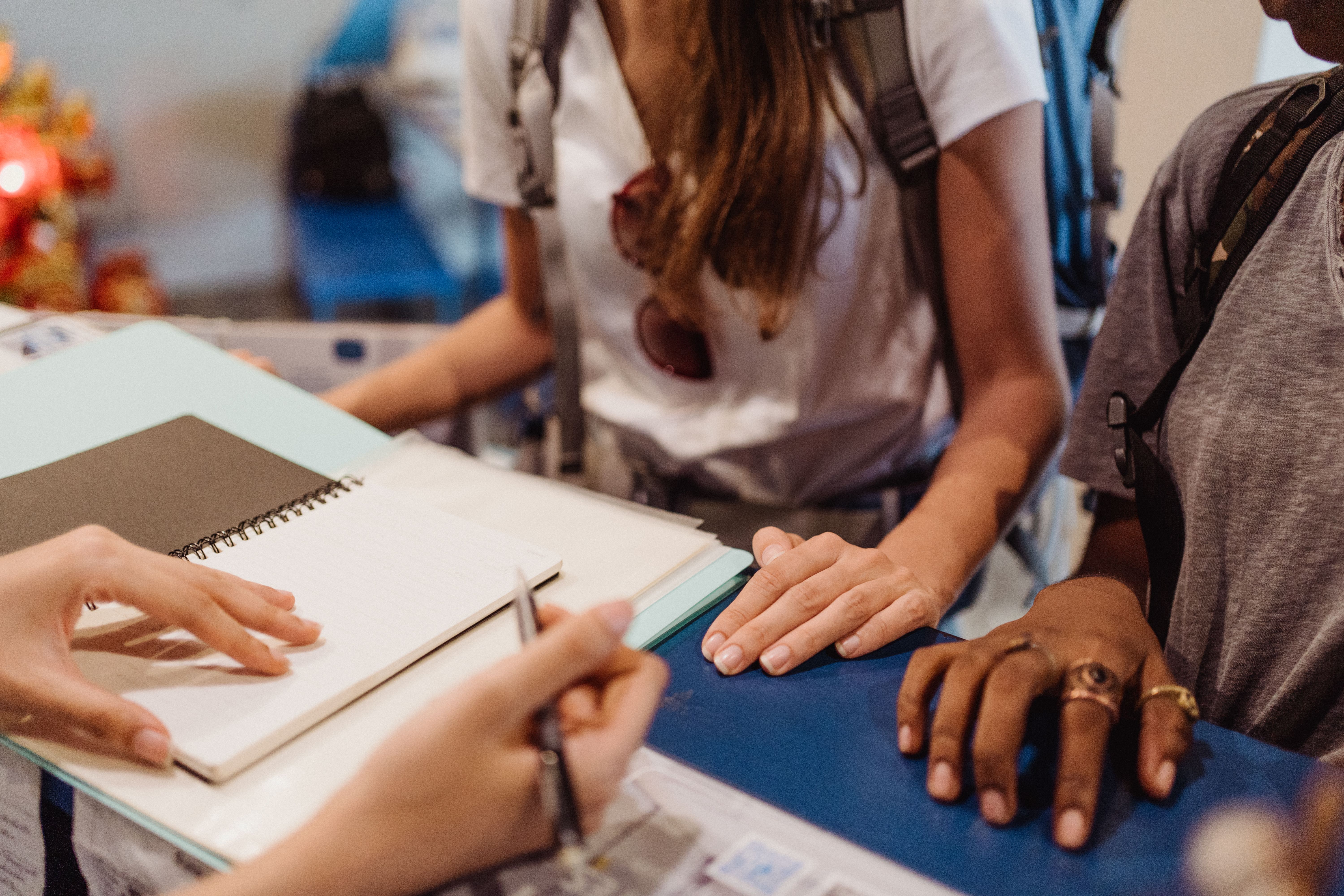 Quy trình xin visa diễn ra như thế nào? Two people with backpacks stand at a reception desk with a staff member holding a pen and notebook, discussing or registering.