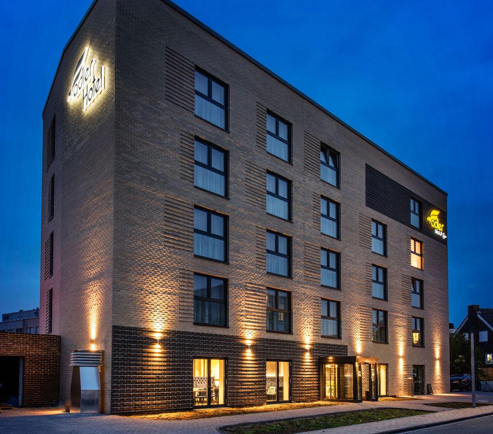 Mehrstöckige, moderne Hotel-Fassade aus Klinkerstein bei Dämmerung, stimmungsvoll inszeniert durch vertikal strahlende LED-Fassadenbeleuchtung und beleuchtete Logos.