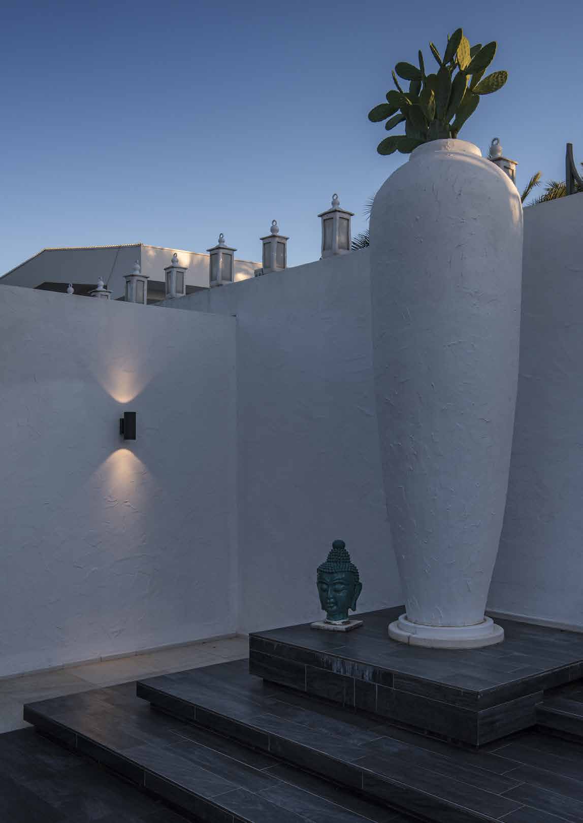 Minimalistische weiße Wand mit der Wandleuchte HELIA PRO, große weiße Vase mit Kaktus auf einem dunklen Sockel, daneben ein kleiner Buddha-Kopf, heller Himmel in der Dämmerung.