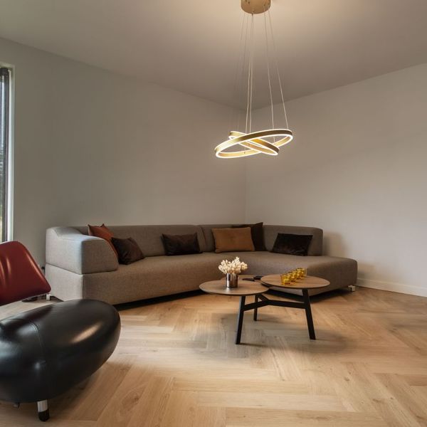 Modernes Wohnzimmer mit grauem Ecksofa, einzigartigem schwarzen Sessel, rundem Couchtisch und der ONE DOUBLE Pendelleuchte in Gold.