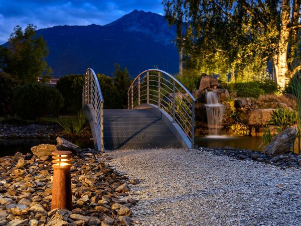 Eine kleine Fußgängerbrücke mit Metallgeländer überspannt einen ruhigen Teich, der von der RUSTY® 40 beleuchtet wird, während im Hintergrund in der Abenddämmerung Berge und Bäume zu sehen sind.