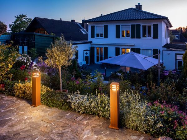 Ein gemütliches Vorstadthaus in der Abenddämmerung, mit einem von RUSTY® Rostleuchten beleuchteten Gartenweg, viel Grün und einem Sonnenschirm unter dem Abendhimmel.